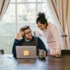First Time Homebuyer Webinar Available On-Demand A man and woman lean over an open laptop with a yellow post-it note stuck to the outside lid at the dining room table with drinks in hand
